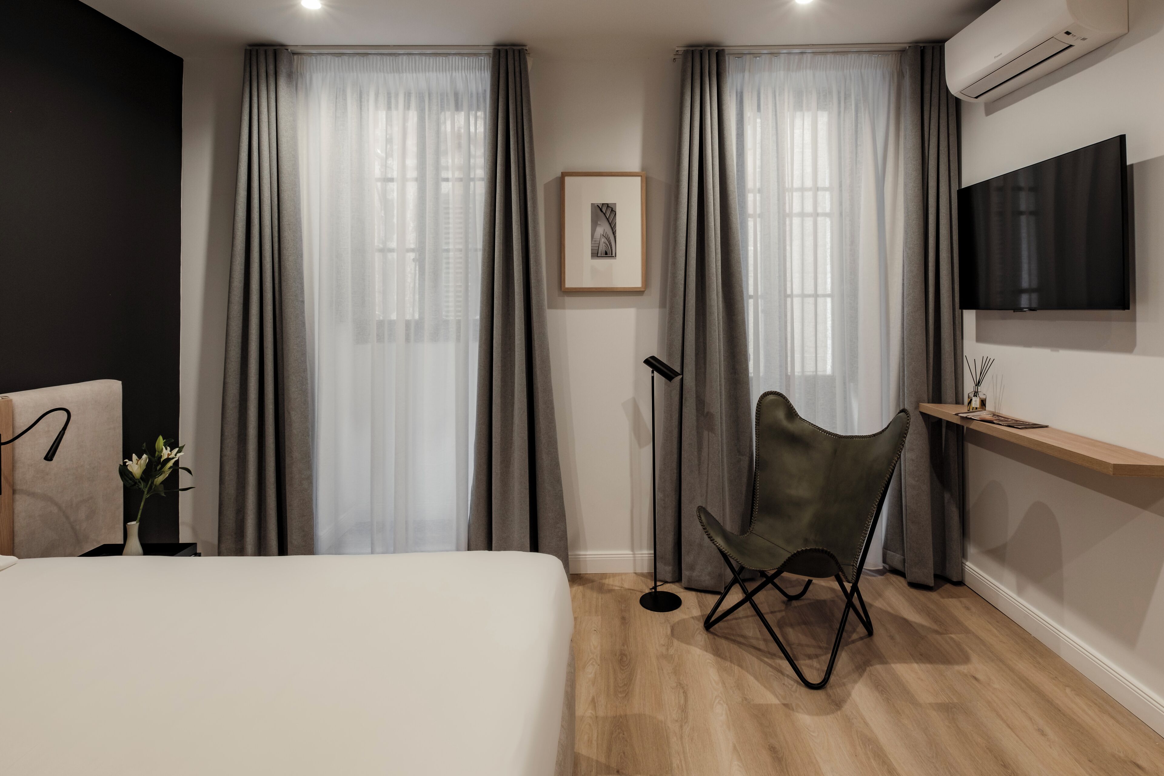 A modern hotel room with a bed, a desk with a chair, a mounted TV, and a large window with curtains.