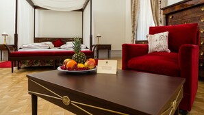 Una cama con dosel, con cabecera tapizada en rojo, un sillón de terciopelo rojo con un cojín, una mesita de noche de madera con una lámpara, y una mesa de centro de madera con un cuenco de fruta y una tarjeta de presentación.