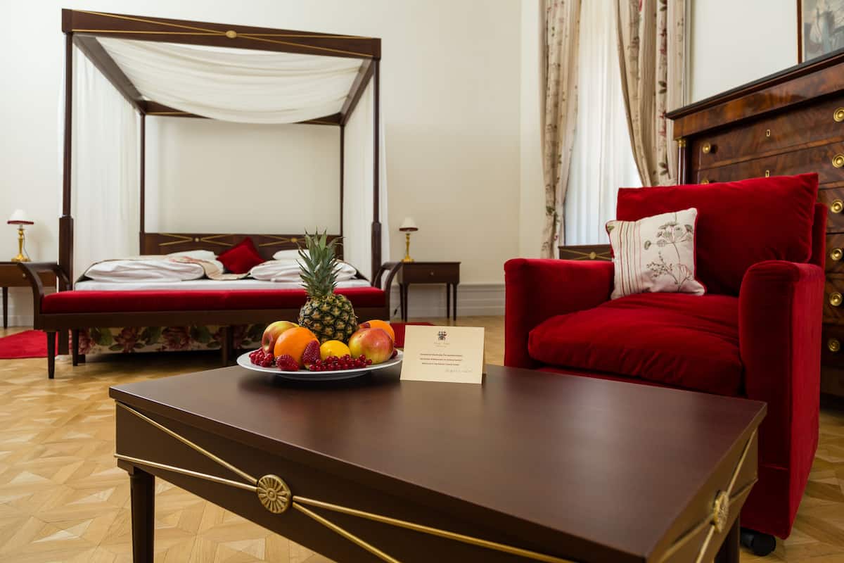 Una cama con dosel, con cabecera tapizada en rojo, un sillón de terciopelo rojo con un cojín, una mesita de noche de madera con una lámpara, y una mesa de centro de madera con un cuenco de fruta y una tarjeta de presentación.