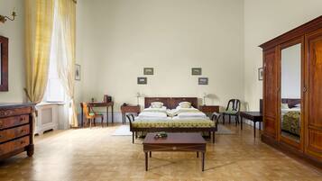 A bedroom with a wooden floor, a bed with green bedding, a wooden dresser, a desk with a chair, and a large mirror.