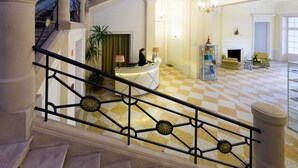 Un vestíbulo de hotel con un mostrador de recepción, una mesa de registro, una escalera con barandilla y un suelo a cuadros.