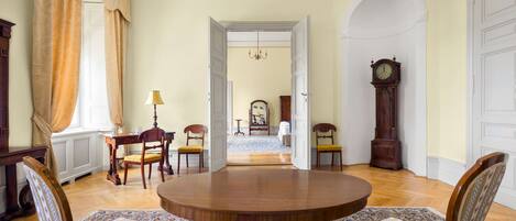 A dining area with a round table, two chairs, a patterned rug, and a view into another room with a chandelier and a clock.