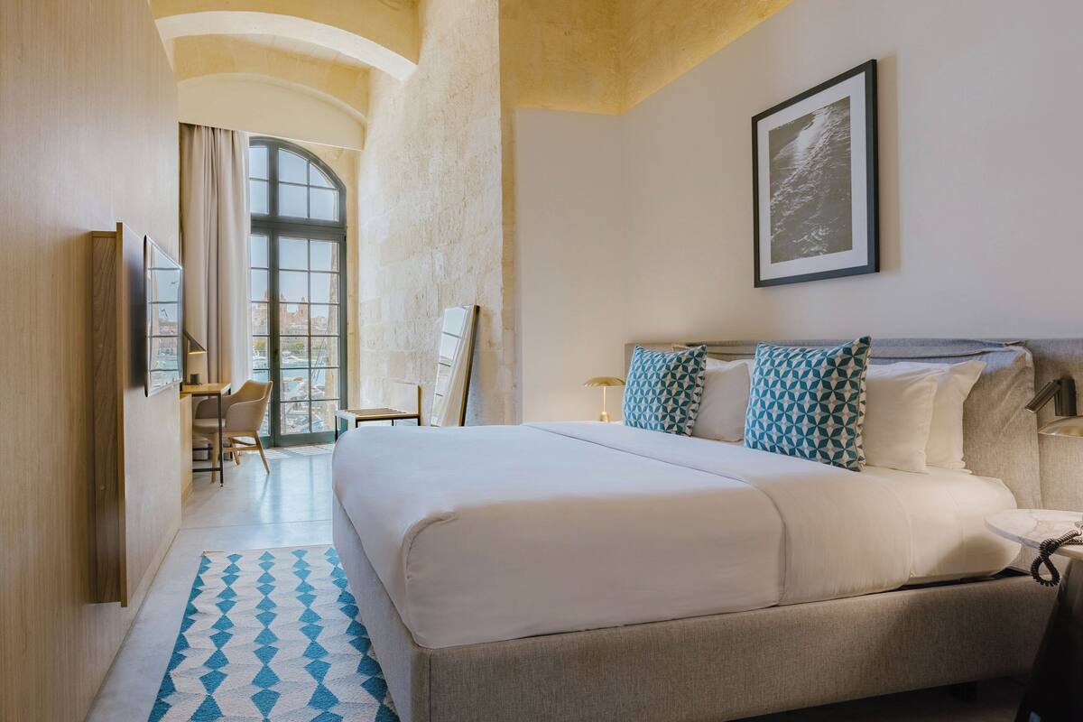 A modern hotel room with a large bed, a desk, and a chair. There is a window with curtains, a framed picture on the wall, and a patterned rug on the floor.