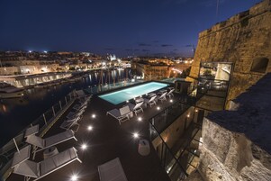 Piscina infinita no terraço com espreguiçadeiras, com vista para um porto com barcos e luzes da cidade.