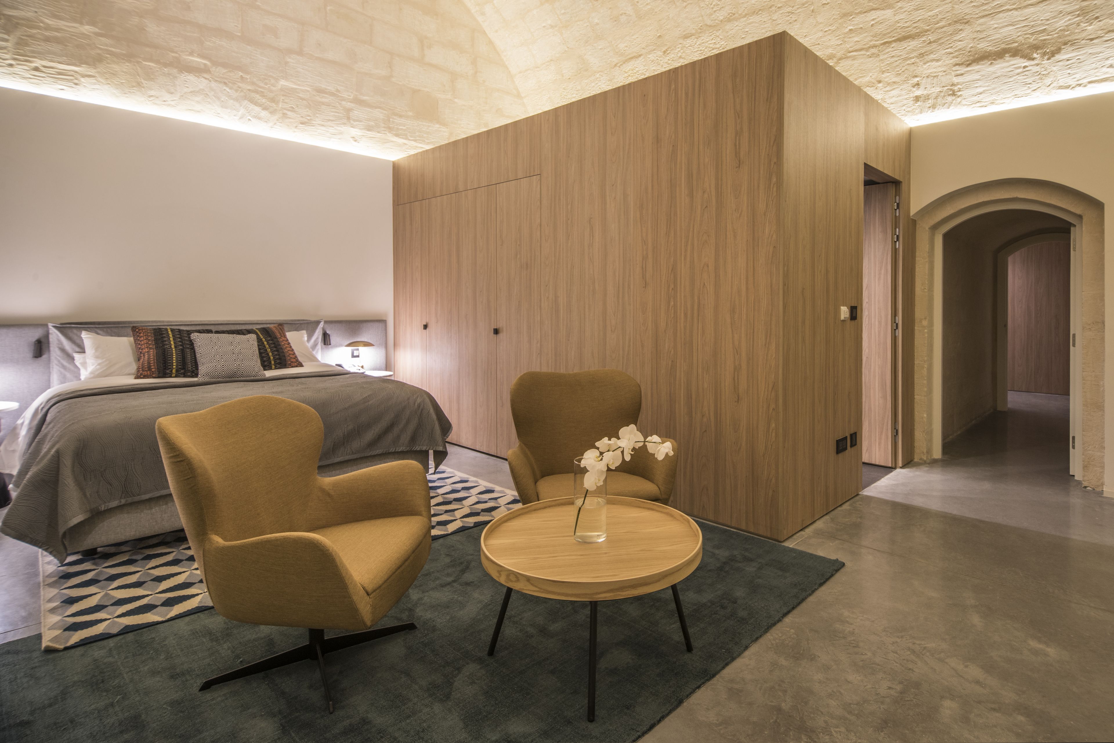 A modern hotel room with a bed, two armchairs, a small wooden table, and a vase with flowers.