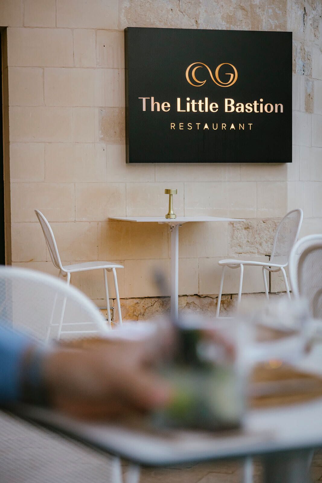 Un letrero de restaurante con logotipo y nombre, sillas blancas y una mesa con una bebida.