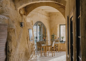 A stone-walled corridor with an arched ceiling, wooden chairs, a table, and a potted plant.