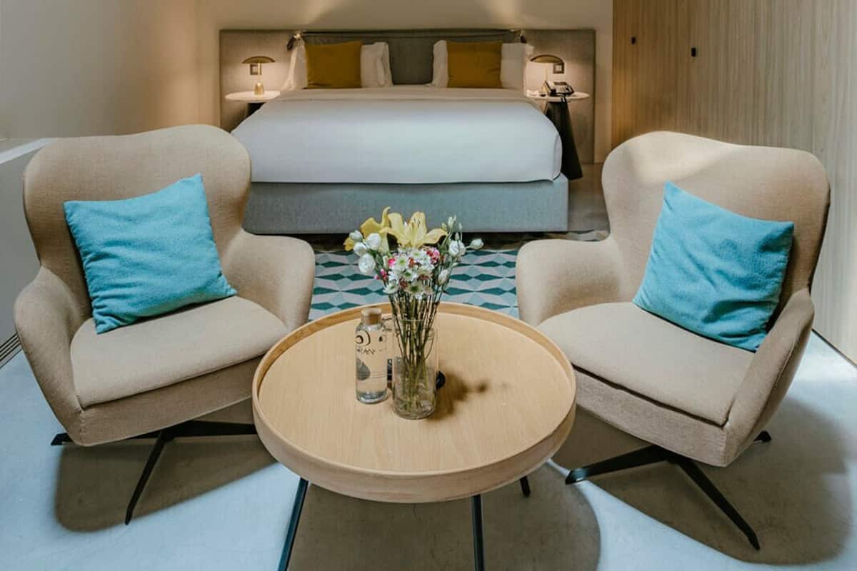 A modern hotel room with a large bed, two armchairs, a round table, and a vase of flowers.