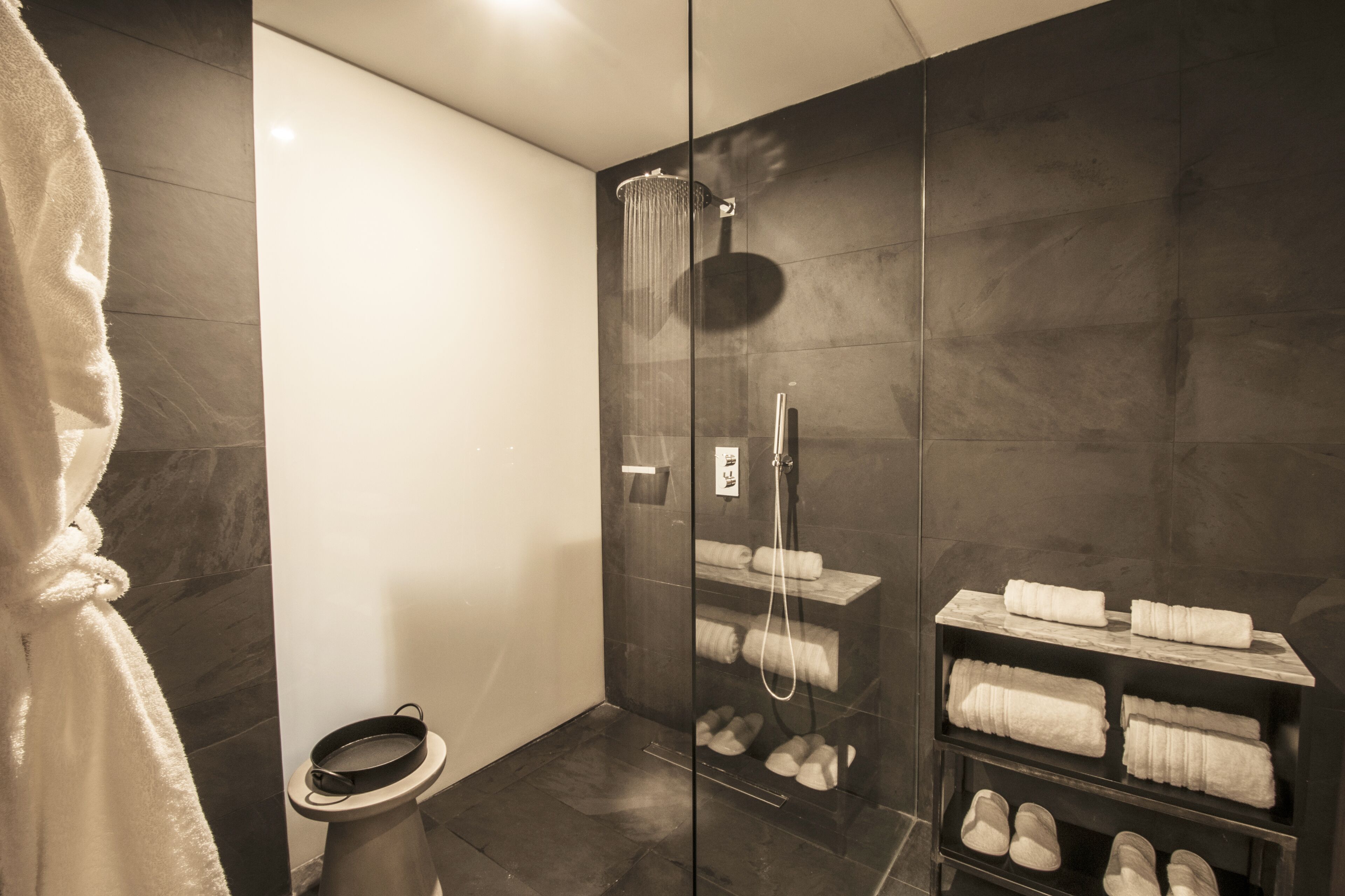 A modern bathroom with a glass-enclosed shower, a toilet, and shelves with neatly folded towels and slippers.