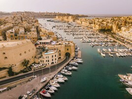 Uma marina com vários barcos atracados, rodeada por edifícios históricos e uma fortificação imponente.