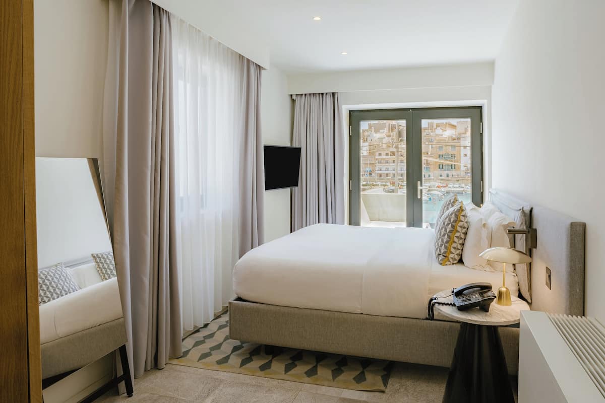 A hotel room with a large bed, a bedside table with a lamp, a telephone, and a TV mounted on the wall.