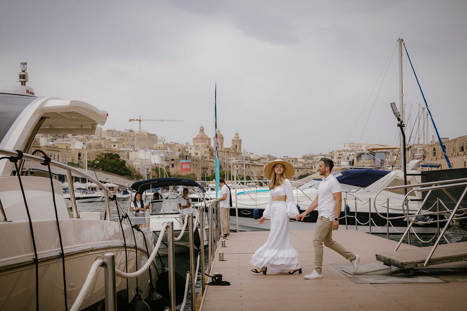 Um casal caminhando em um cais com barcos e um horizonte urbano ao fundo.