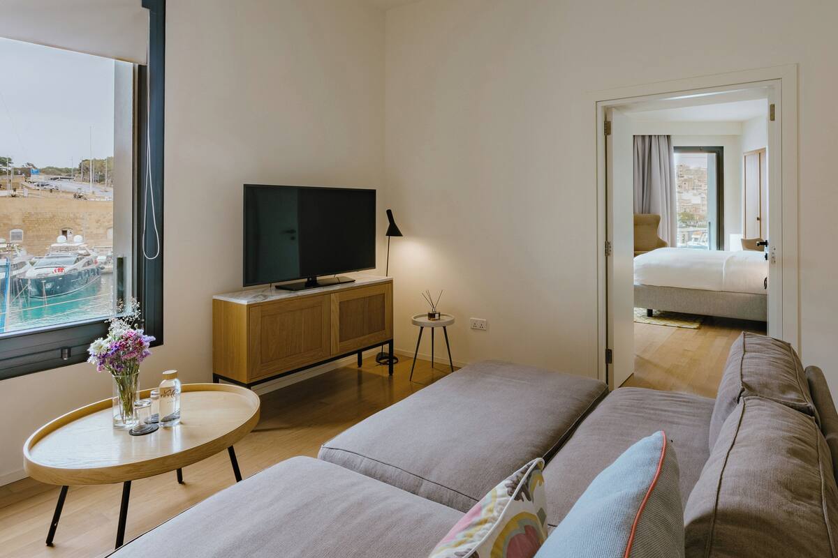 A modern hotel room with a large window, a flat-screen TV, a sofa, a small round table, and a bed with a view of the city.