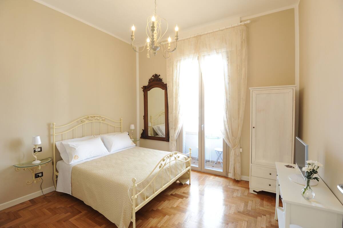 Ein Schlafzimmer mit Bett, Nachttisch, Lampe, Spiegel, Kleiderschrank und Schreibtisch mit Bildschirm und Blumen.