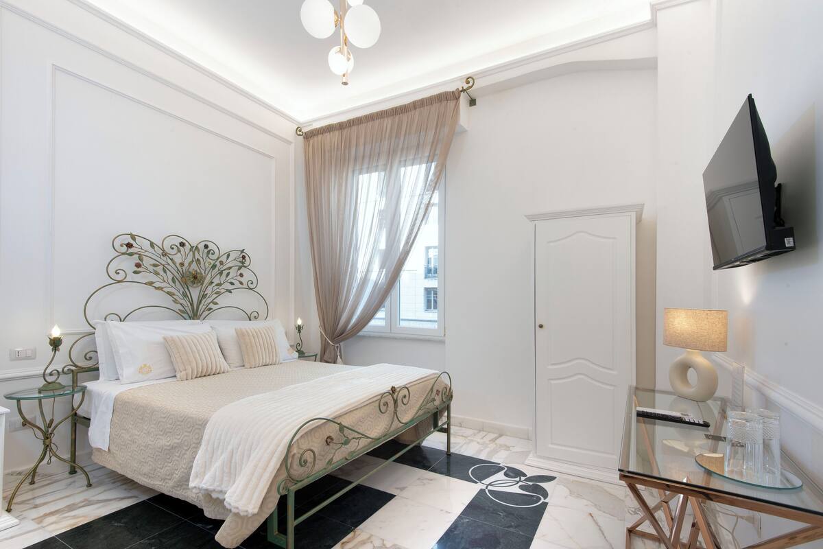 Ein Schlafzimmer mit Bett, Nachttisch, Glastisch, Fernseher und einem Fenster mit Vorhängen.