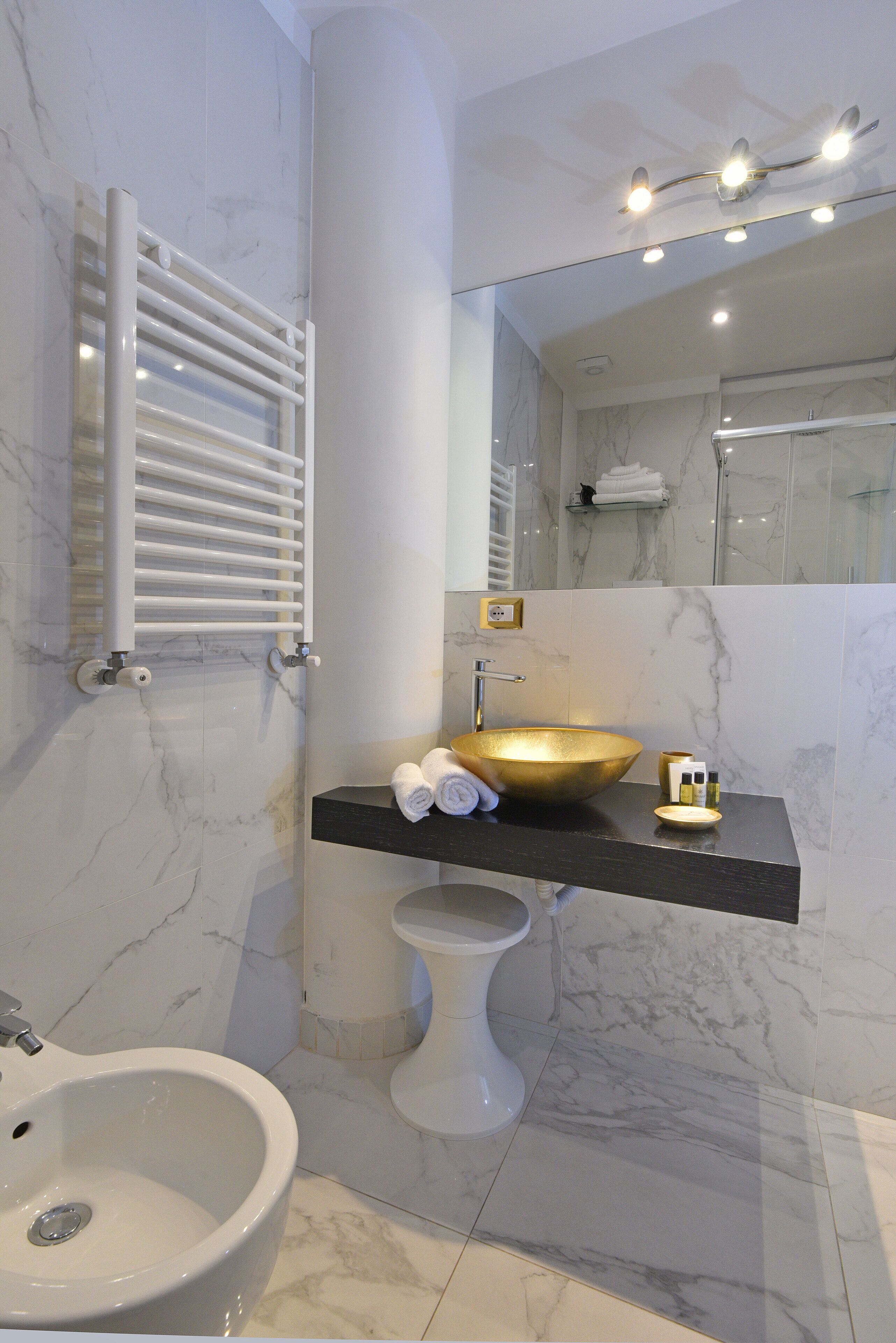 A modern bathroom with a marble floor, a gold sink, a white pedestal sink, a white bidet, and a towel rack.