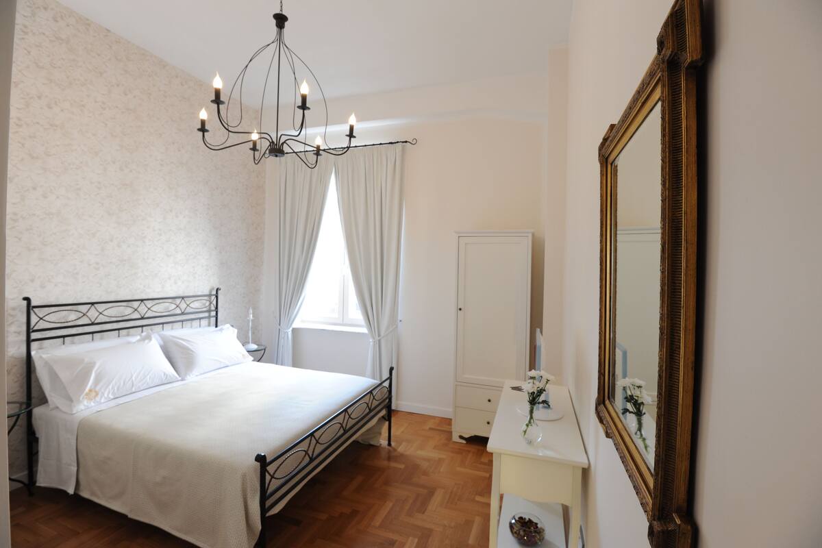 Ein Schlafzimmer mit einem Bett, einer Kandelaber, einem Spiegel und einem Fenster mit Vorhängen.