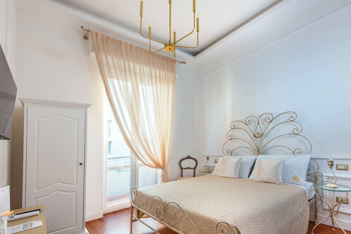 Ein Schlafzimmer mit Bett, Vorhängen, einer Pendelleuchte, einem Nachttisch und einem Beistelltisch.