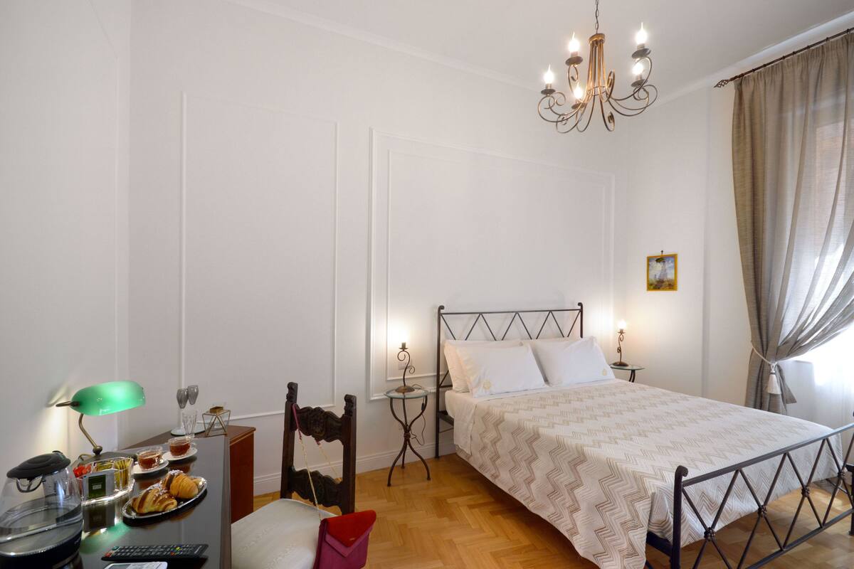 Ein ordentlich eingerichtetes Schlafzimmer mit Bett, Nachttischen, einem Schreibtisch mit Lampe und einem Sessel.