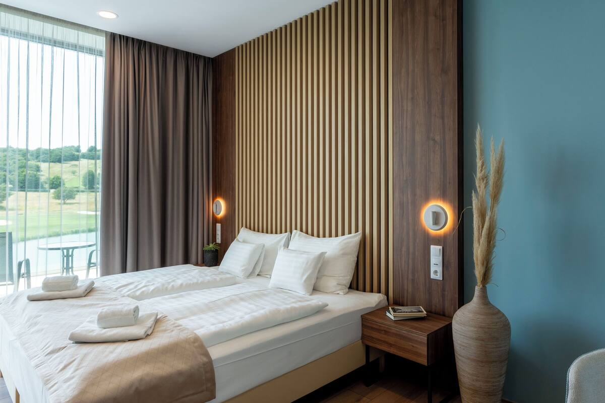 Habitación de hotel moderna con una cama grande, dos mesitas de noche, vistas al exterior y una pared de acento de madera.