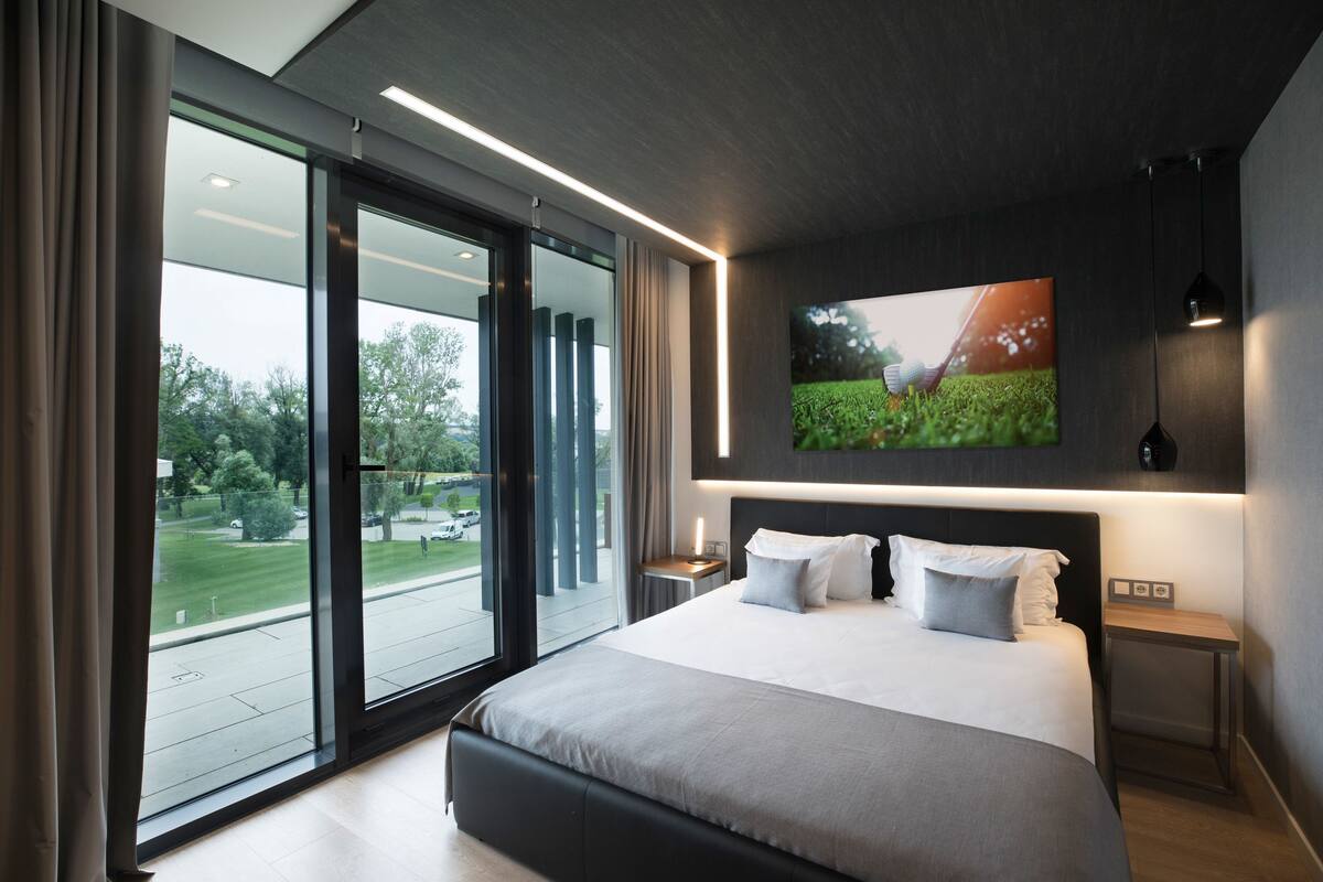 Habitación de hotel moderna con una cama grande, un televisor montado en la pared y un ventanal corredero que da a un balcón con vistas al exterior.