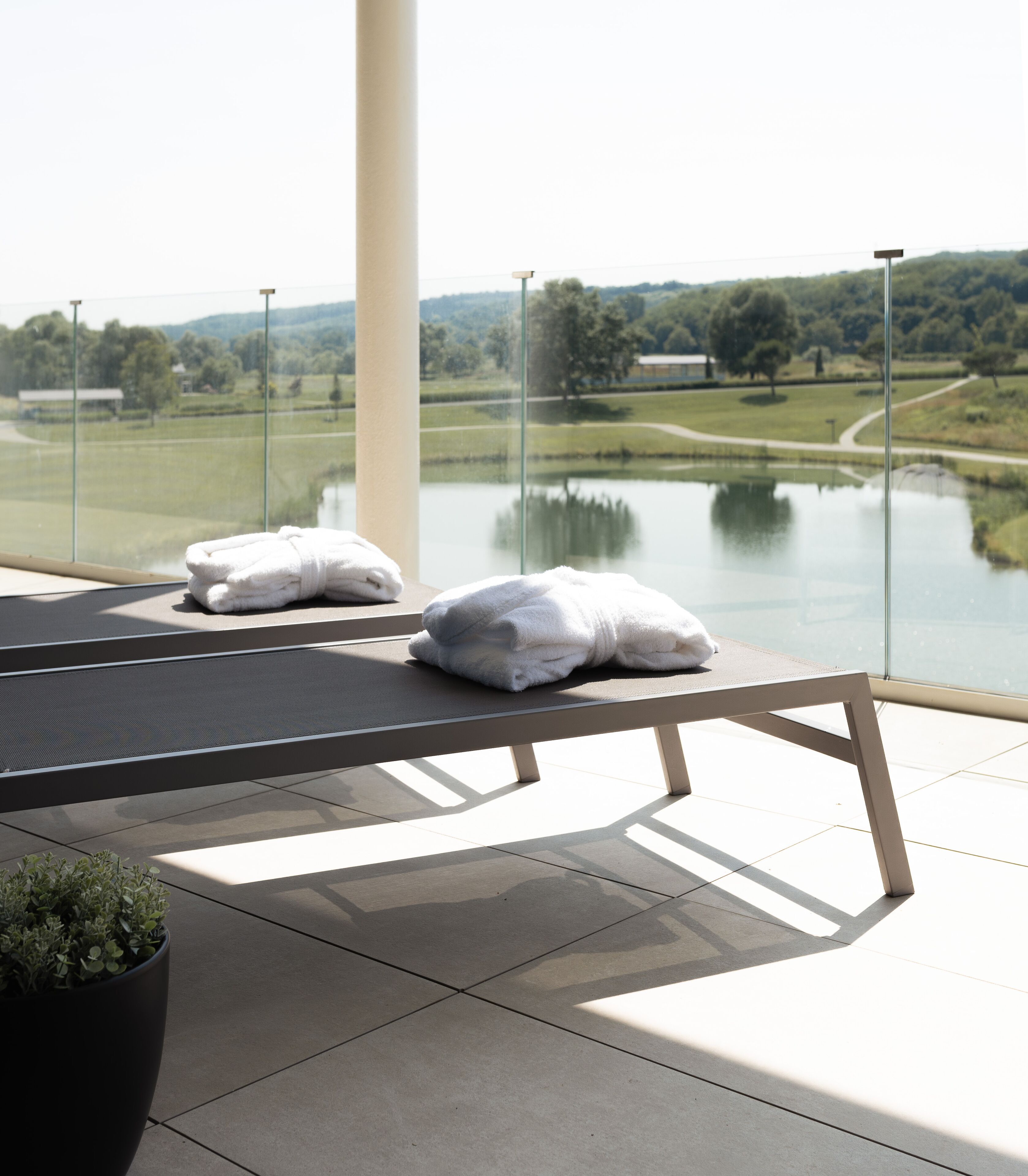 A balcony with a view of a lake and hills, two white towels on a bench, and a potted plant.