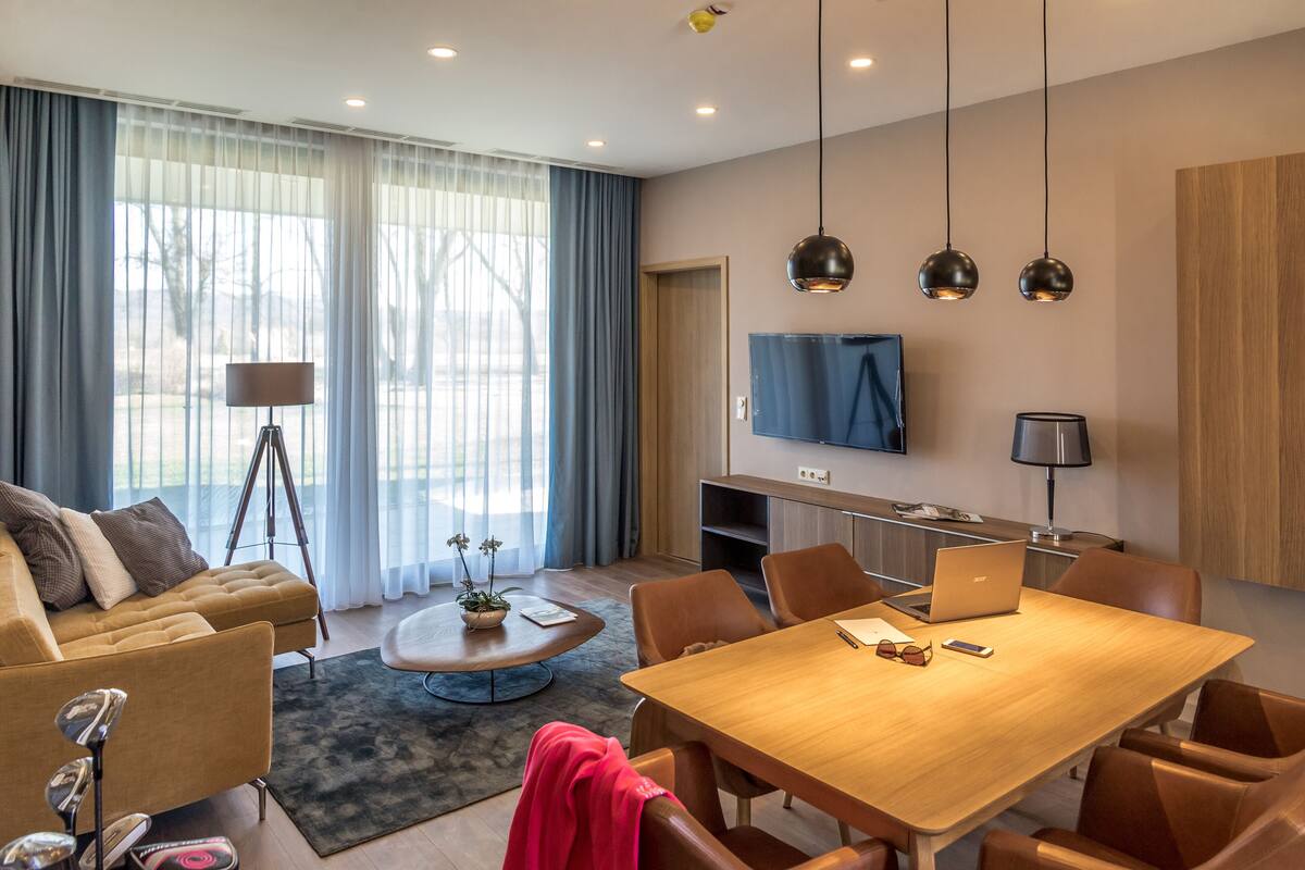 Una sala moderna con sofá, una mesa de centro y una mesa de comedor. Hay un televisor montado en la pared y un ordenador portátil sobre la mesa.