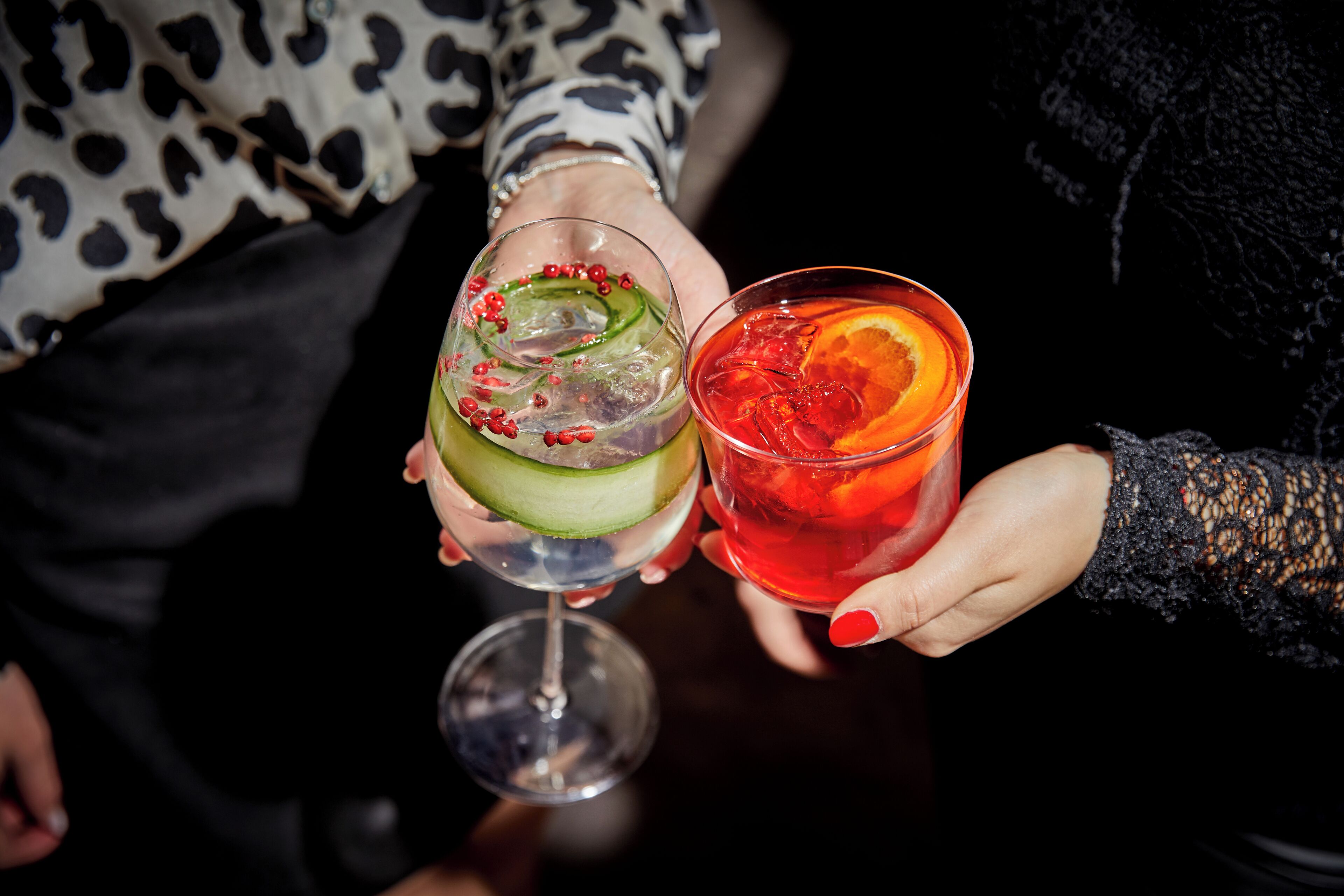 Zwei Personen mit Cocktailgläsern, eine hält ein grünes Getränk mit Gurke und Granatapfelkernen, die andere ein rotes Getränk mit Orangenscheibe.