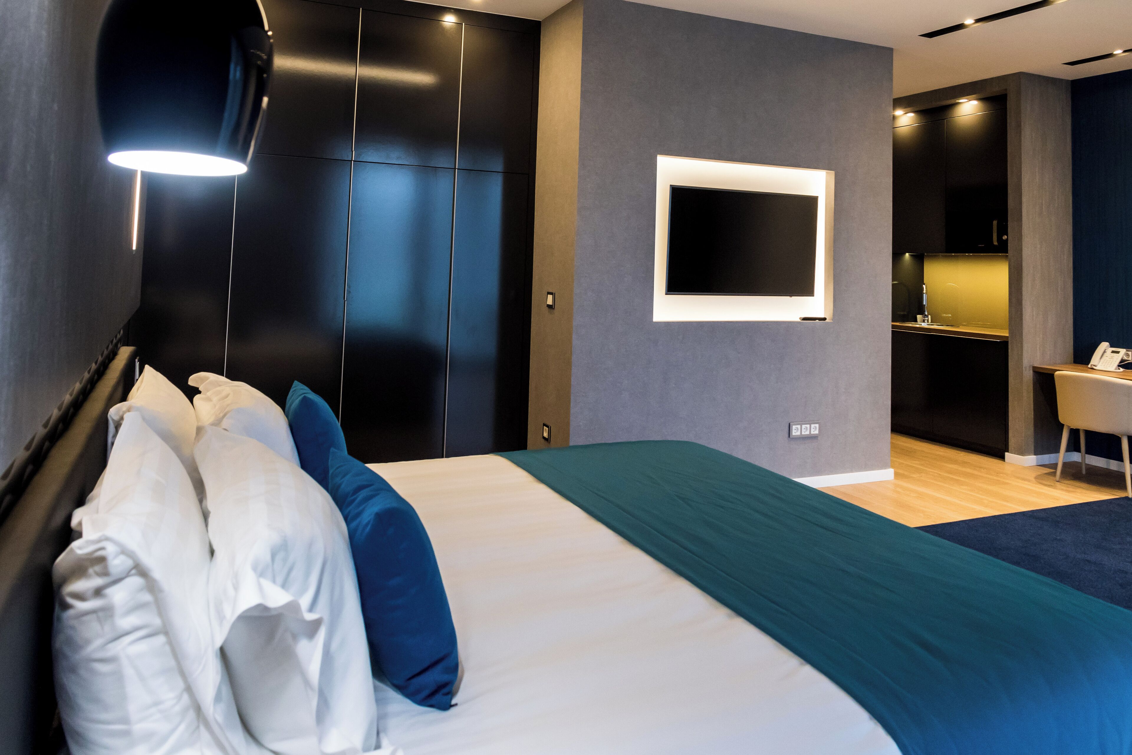 A modern hotel room with a large bed, dark walls, a TV mounted on the wall, and a small desk area.