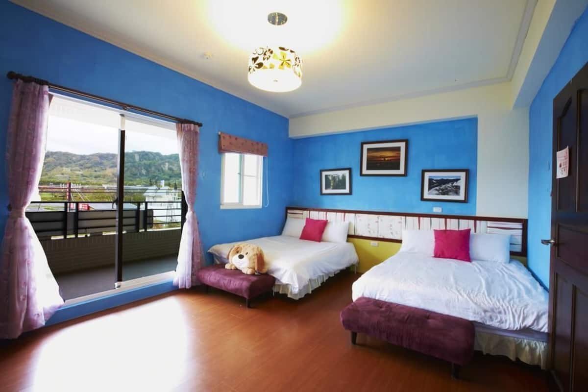 A room with two beds, a large window with curtains, a ceiling light fixture, and framed pictures on the wall.