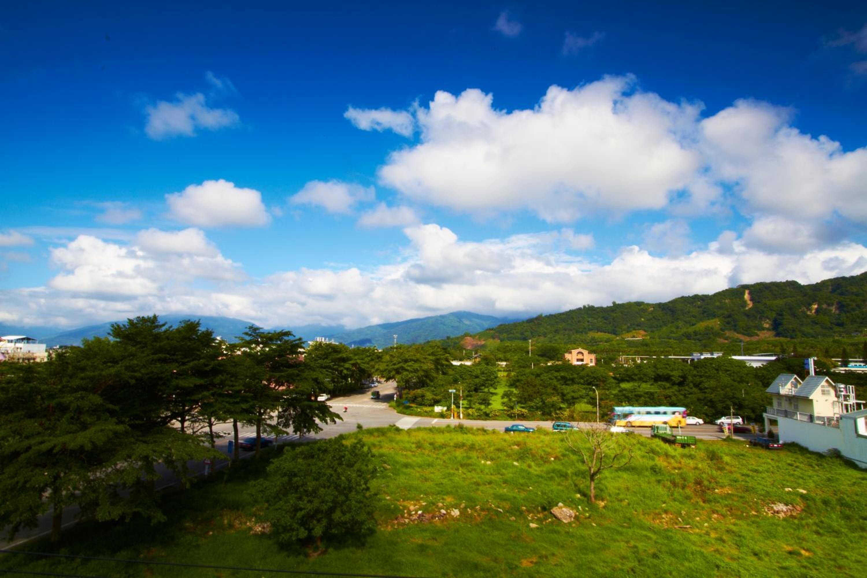 A scenic view of a town with a road leading to a hilltop building, surrounded by greenery and mountains in the background.