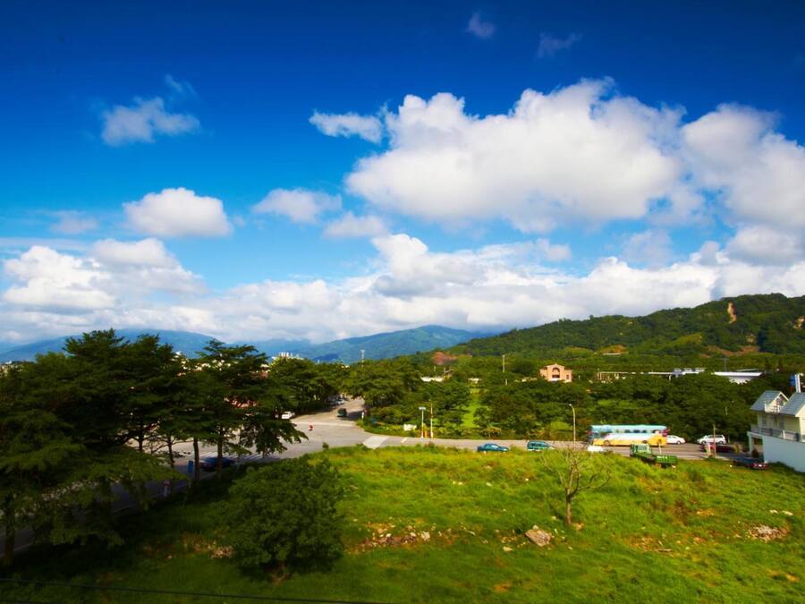 A scenic view of a town with a road leading to a hilltop building, surrounded by greenery and mountains in the background.