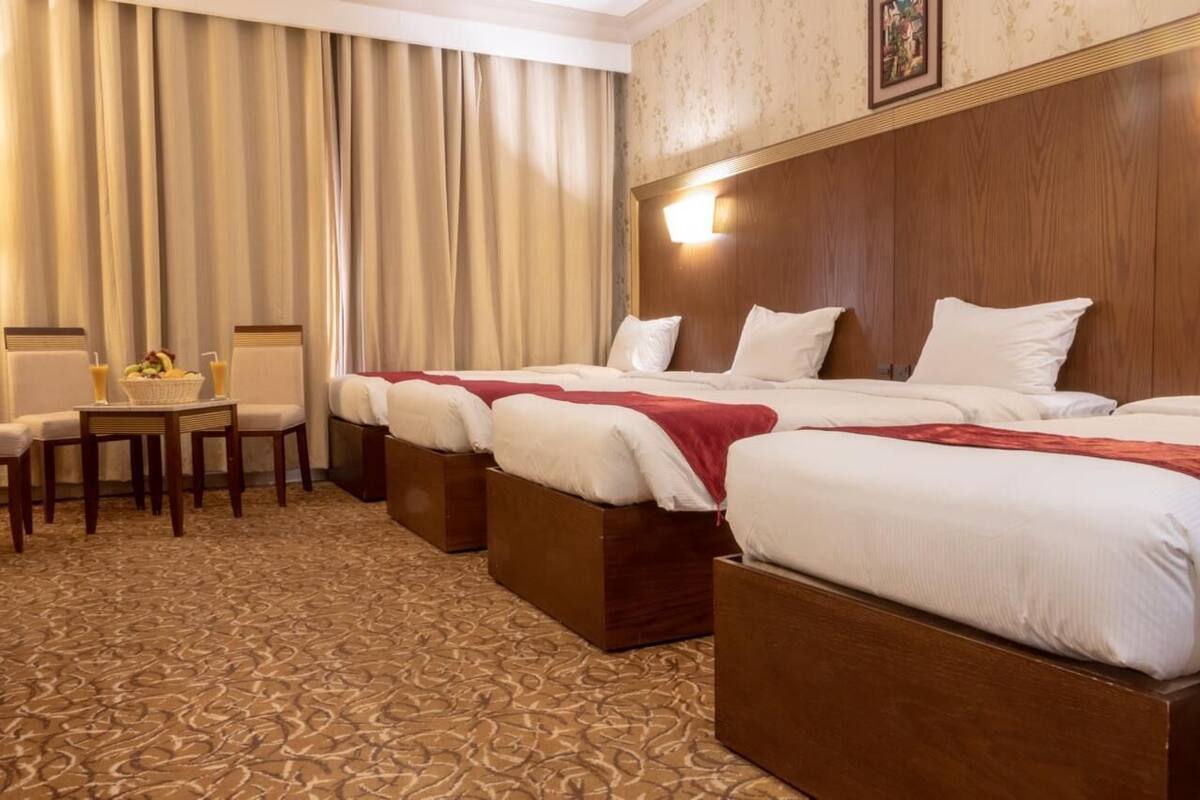 A hotel room with three beds, a dining table with chairs, and a small table with a fruit bowl and drinks.