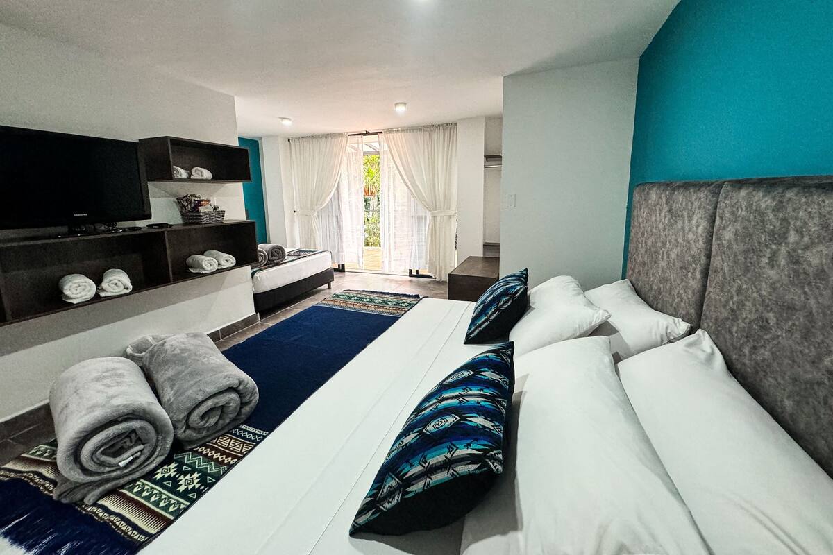 Quarto de hotel moderno com uma cama grande, televisão, sofá e varanda com cortinas.
