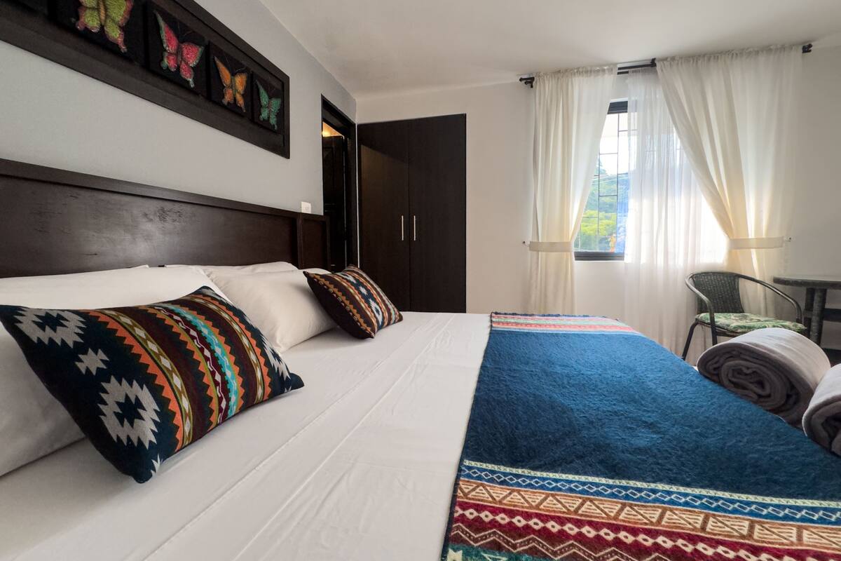 Quarto de hotel com uma cama grande, duas almofadas, cabeceira, janela com cortinas, uma cadeira e uma pequena mesa.