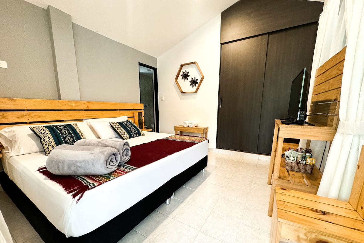Cama bem arrumada com manta vermelha e toalhas dobradas, cabeceira de madeira e mesa de cabeceira de madeira com abajur.