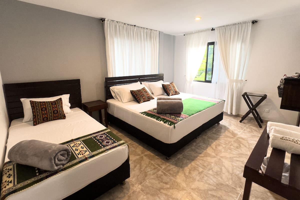 Quarto de hotel com duas camas, uma mesa pequena e uma janela com cortinas.