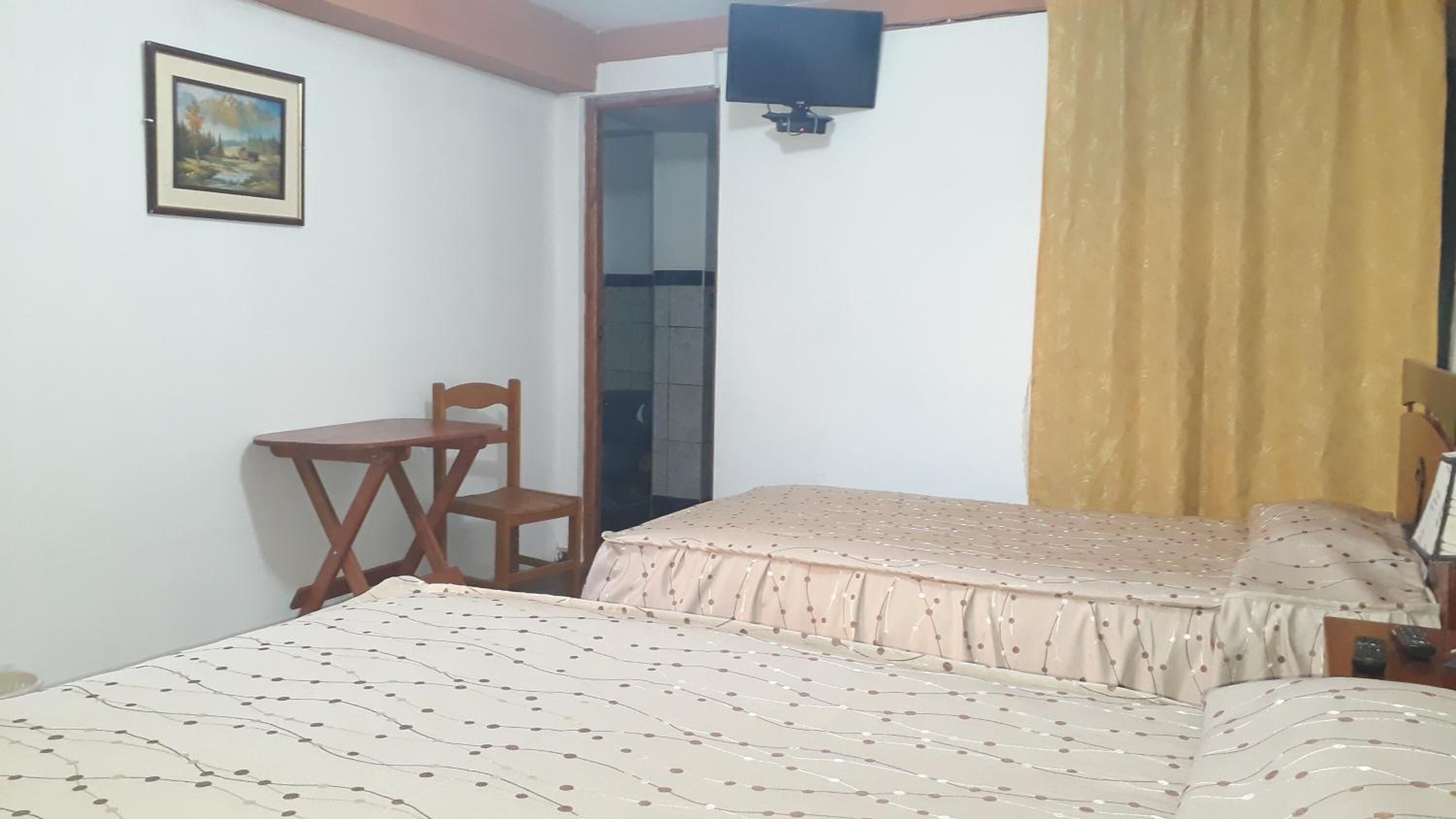 Une chambre à coucher comprenant un lit, une chaise, une petite table, un téléviseur fixé au mur et une fenêtre avec un rideau.