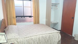 Un dormitorio con una cama, una ventana con cortinas, una puerta y un estante pequeño con ropa doblada.