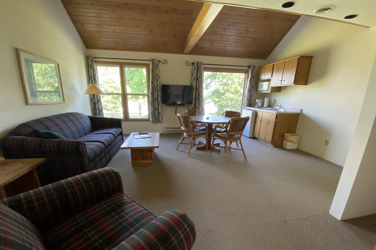 Ein gemütliches Wohnzimmer mit Sofa, Esstisch und Stühlen, Küchenzeile mit Schränken und Mikrowelle sowie großen Fenstern mit Vorhängen.