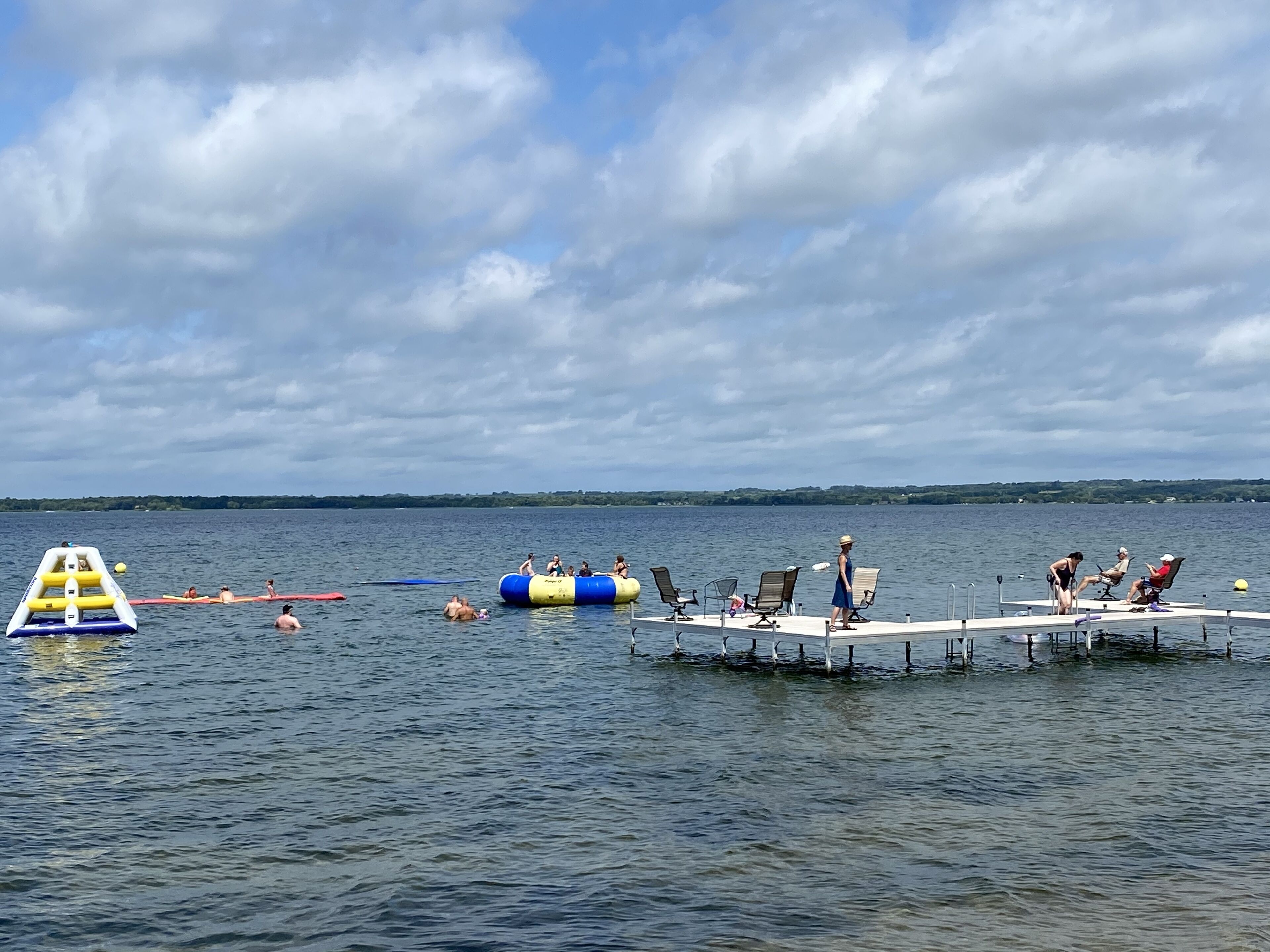 Escena en la orilla de un lago con un muelle, flotador inflable y personas disfrutando de actividades acuáticas.