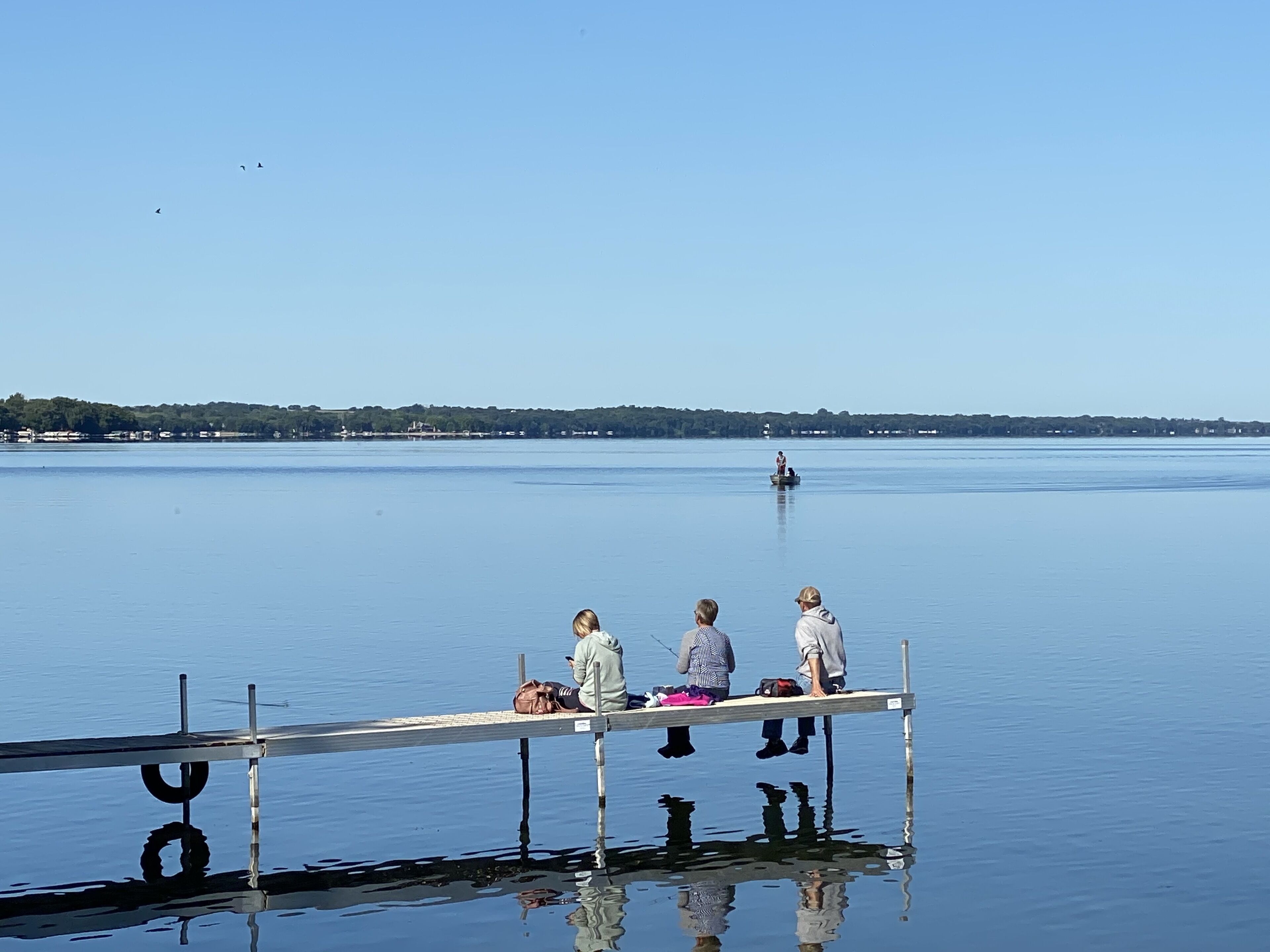 A pier extending into calm water with three people sitting on it, a distant shoreline, and a clear sky.
