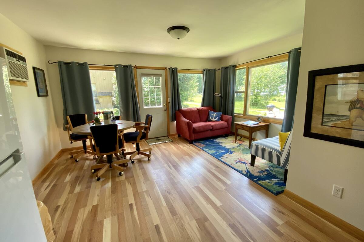 Ein geräumiger Raum mit Holzboden, einem Esstisch mit Stühlen, einem roten Sofa, einem kleinen Beistelltisch und einem Fenster mit Vorhängen.
