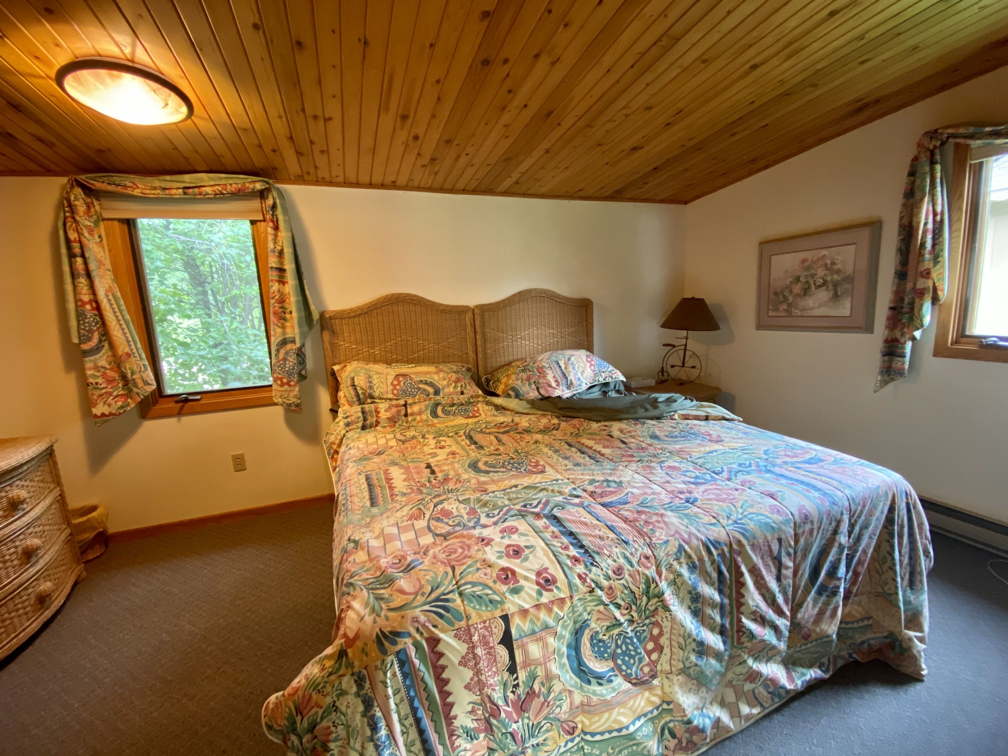 A bedroom with a wooden ceiling, a large bed with a patterned bedspread, a window with curtains, and a nightstand with a lamp.