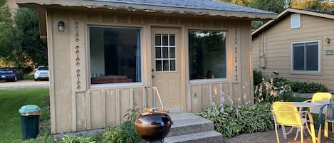Una cabaña de madera pequeña con un porche, una parrilla y un patio con sillas.