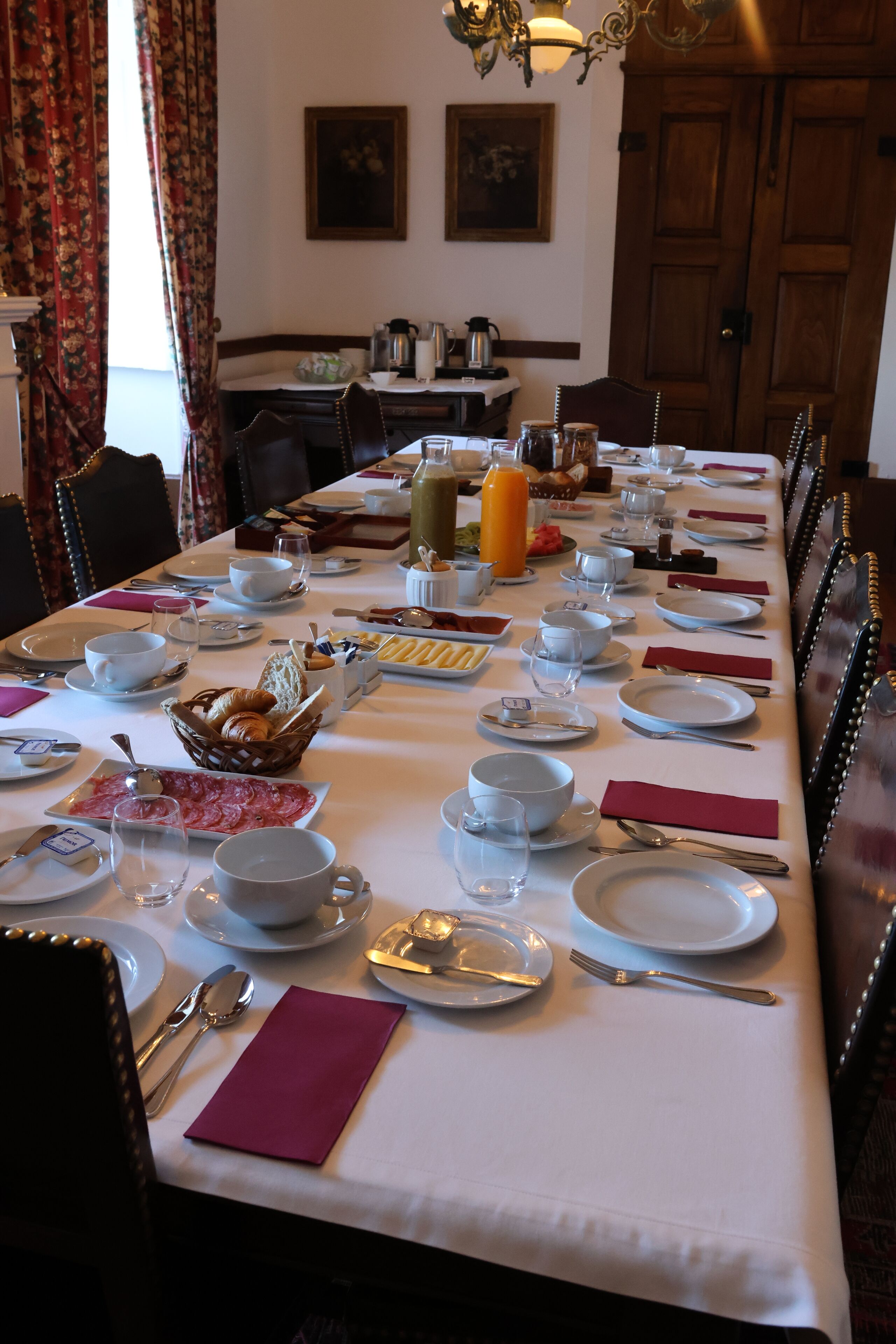 Una larga mesa de comedor dispuesta con platos, tazones, vasos y diversos alimentos, rodeada de sillas.