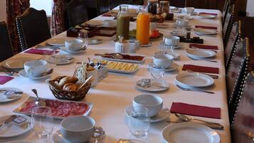 Una larga mesa de comedor dispuesta con platos, tazones, vasos y diversos alimentos, rodeada de sillas.