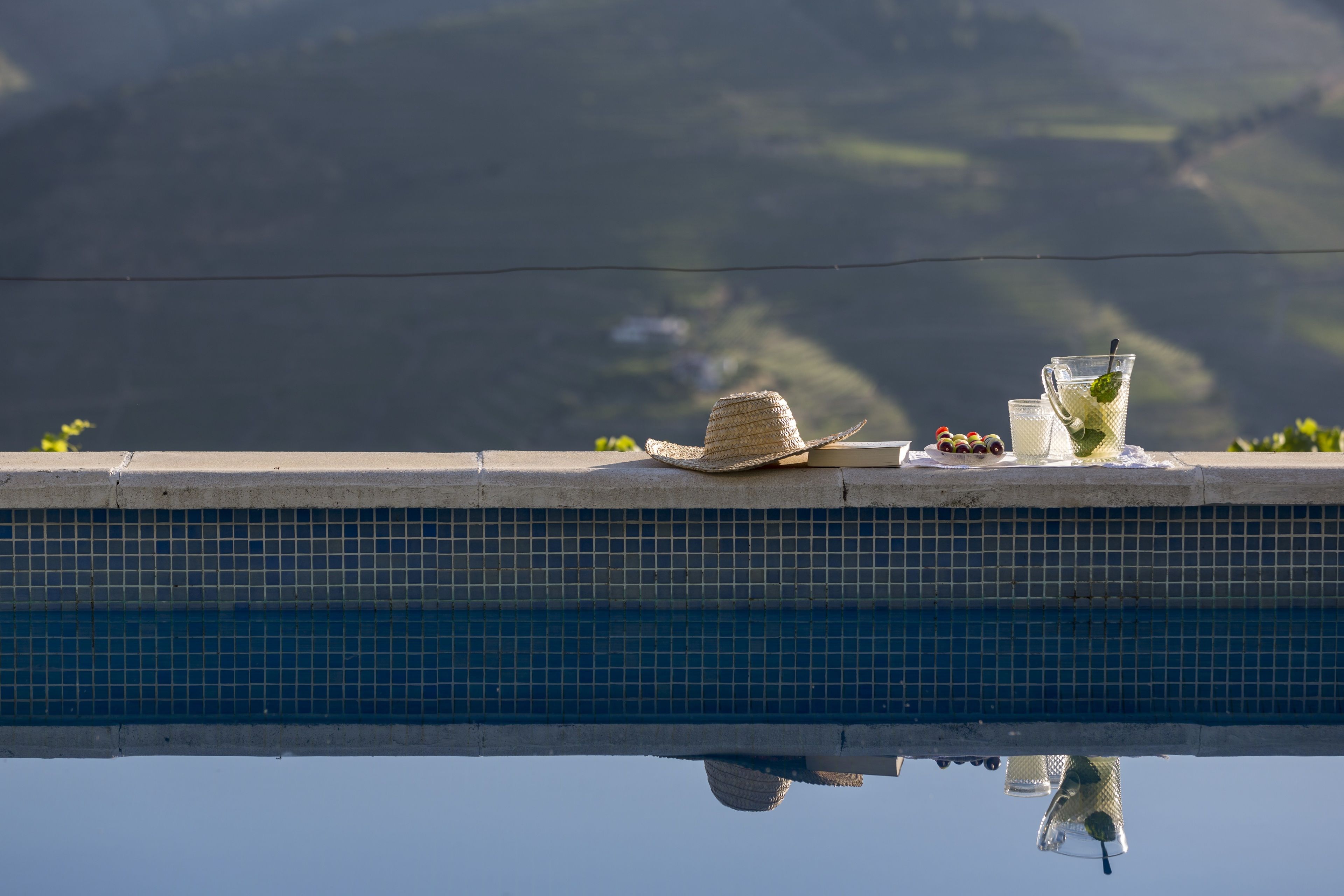 Une vue sur la piscine, avec un chapeau, des lunettes et un verre posé sur le bord.