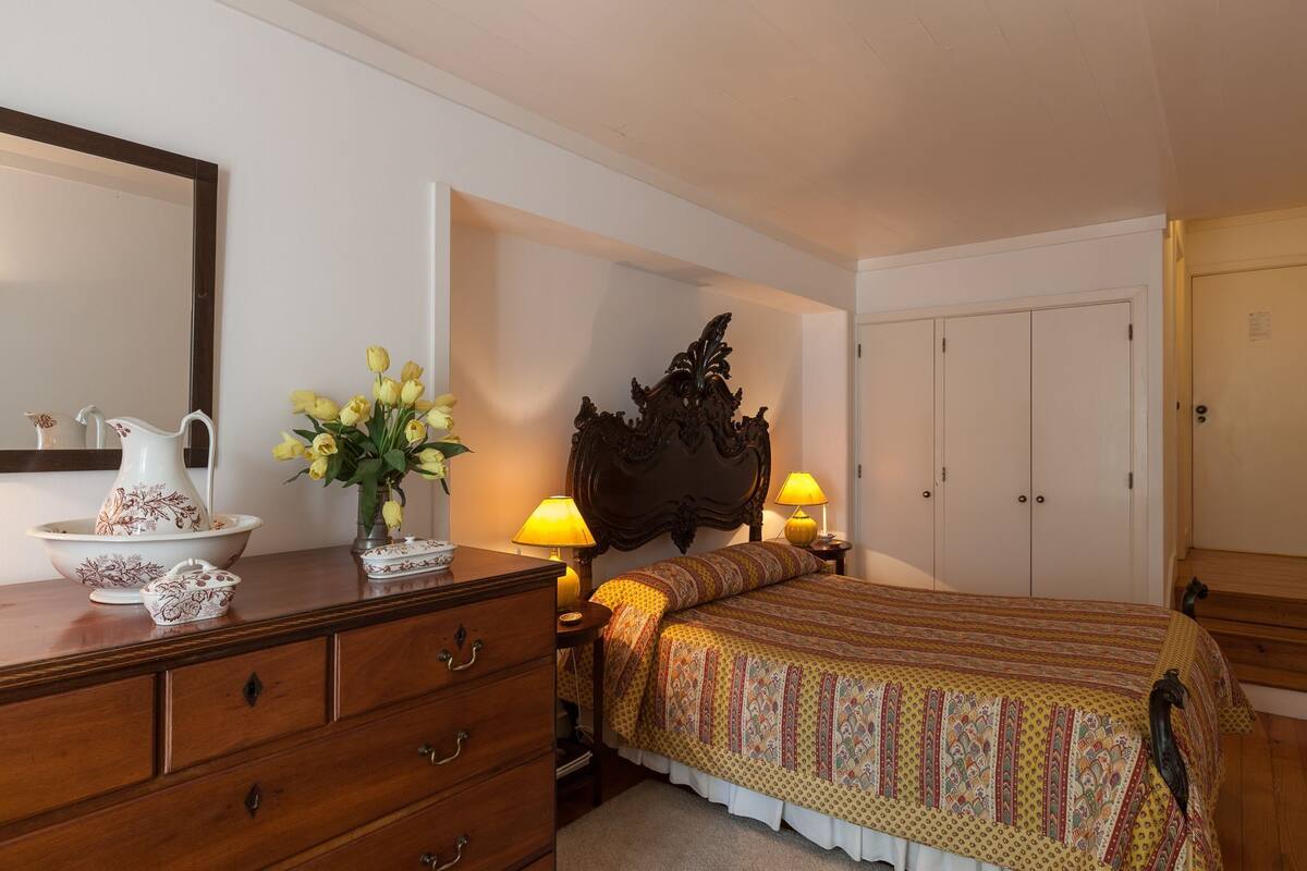 A bedroom with a wooden dresser, a bed with patterned bedding, a nightstand with lamps, and a mirror on the wall.