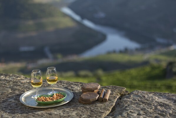 Una vista panorámica de un valle fluvial con una mesa sobre roca, donde se aprecian dos copas de vino blanco, un plato de frutos secos y bloques de madera.